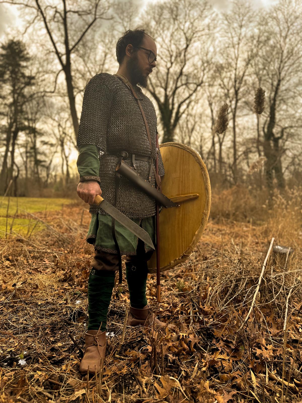 A Viking warrior in a dreary background holding a shield and seax, staring down at the ground
